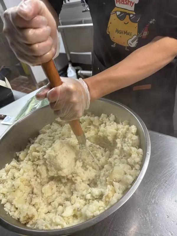 A MashMore crew member in a Mashy apron mashing a big bowl of potatoes